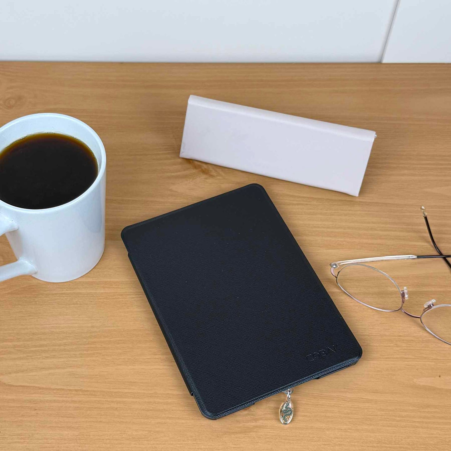 Celestial Snake Kindle Charm USB-C dust plug attached to a black Kindle on a wooden table with coffee and glasses.