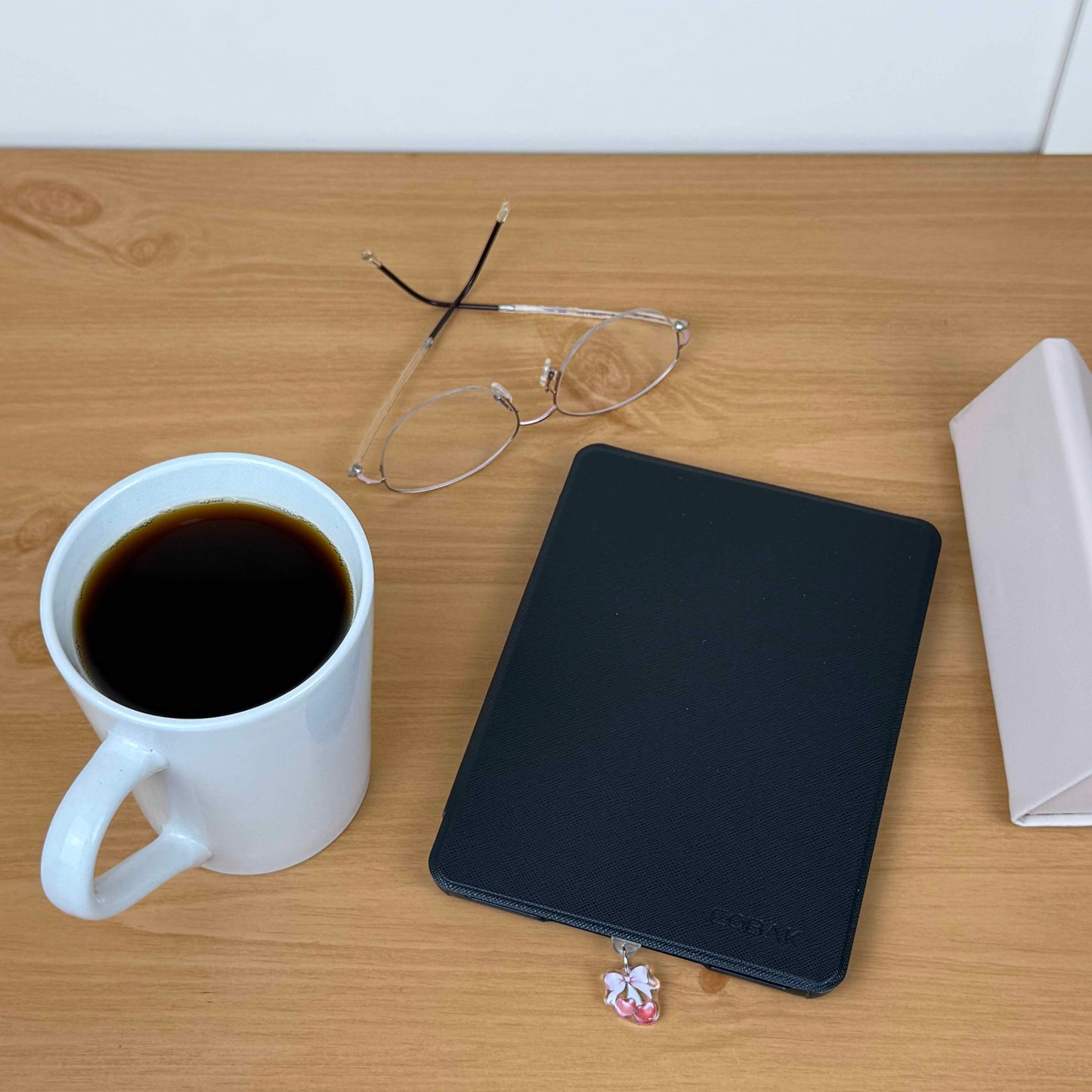 Cherries & Bow Kindle Charm USB-C dust plug attached to a black Kindle on a wooden table with coffee and glasses.