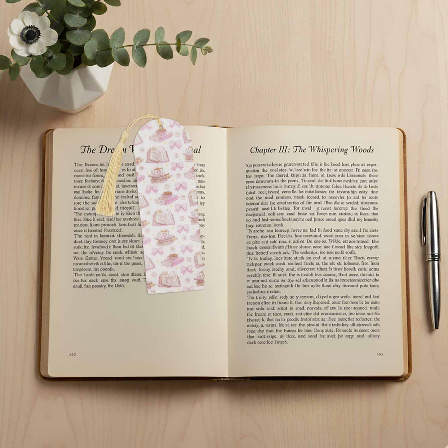 Pink bookmark with books and bows on an open book, with a pen and eucalyptus in the background.