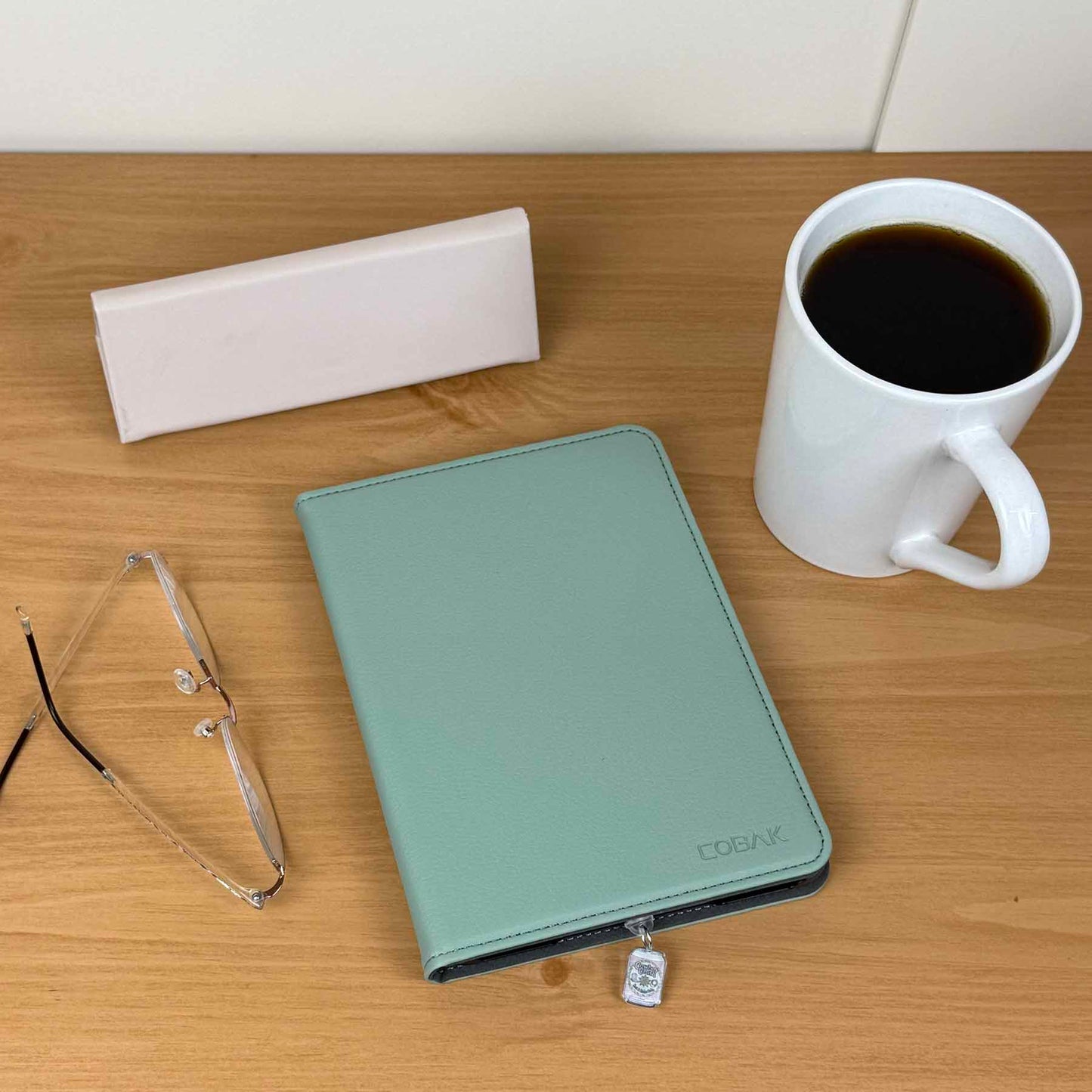 Close-up of a Cowboy Crush Soda Can Kindle Charm attached to a light green Kindle cover on a wooden table, next to a white coffee mug and eyeglasses.