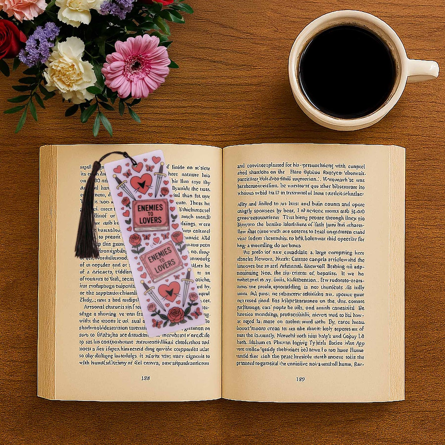 An open book with an "Enemies to Lovers" bookmark, a cup of coffee, and a bouquet of pink and white flowers on a wooden table.