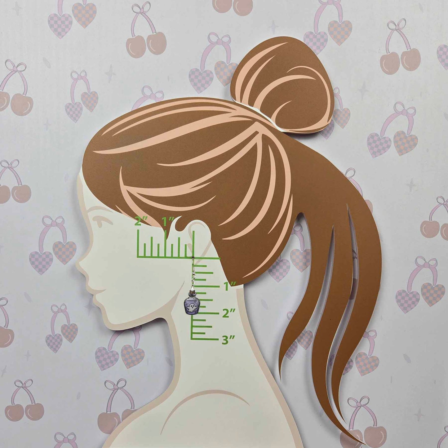 Spooky Ghost Potion Bottle Dangle Earring, purple with white ghost and silver hook, shown on a measurement display against a cherry and heart patterned background.