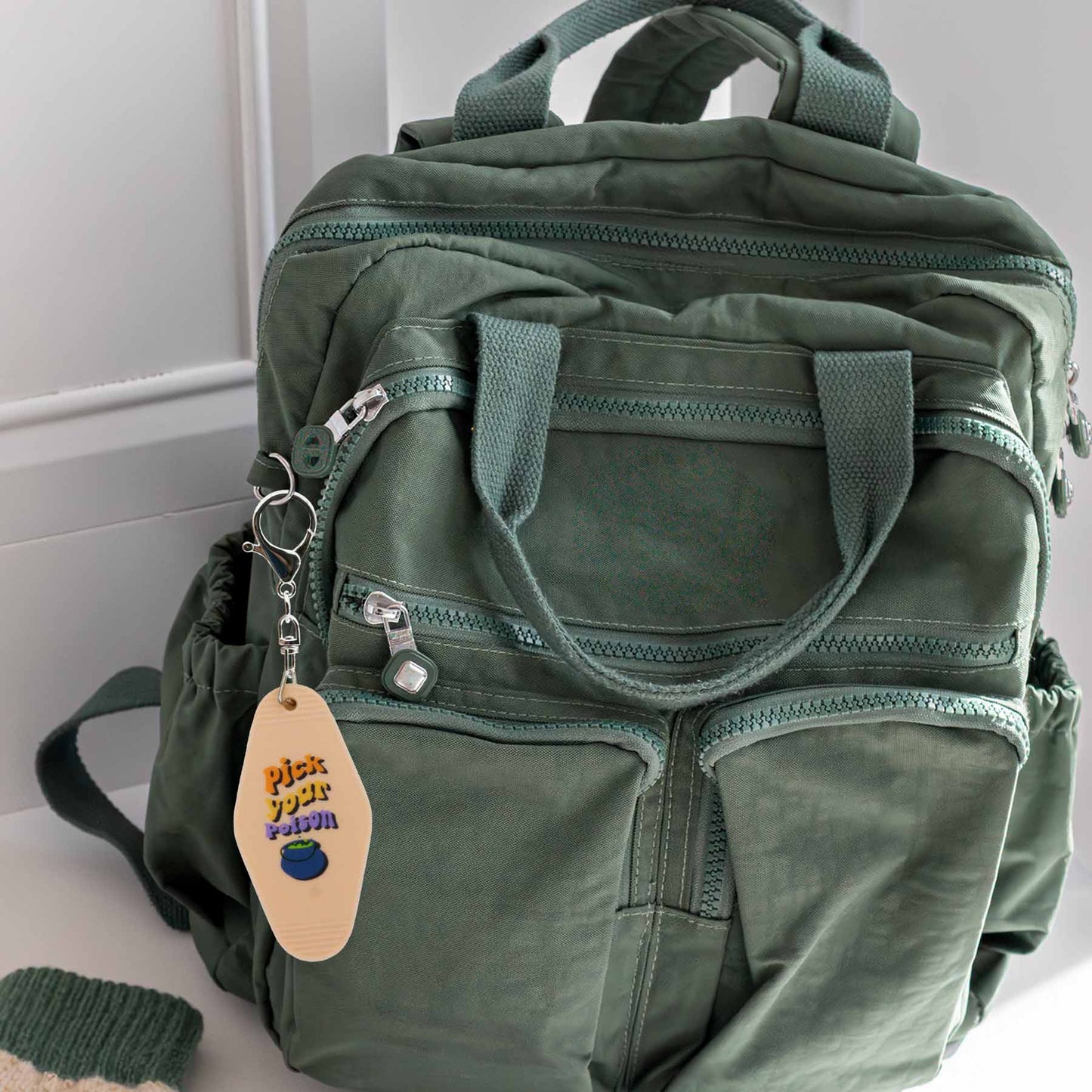 Green military-style backpack with multiple zippered pockets and a beige keychain that says 'Pick Your Poison' with a blue bowl illustration.