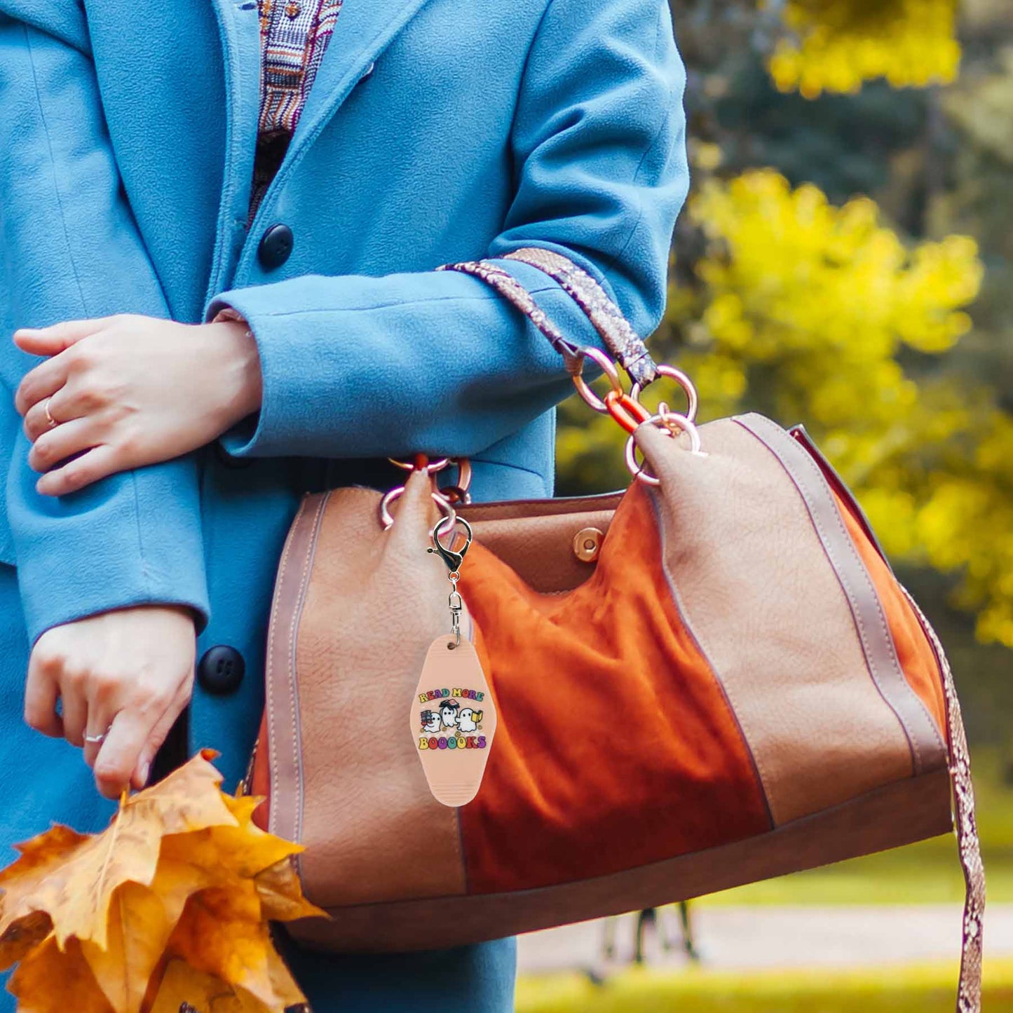 A beige and orange handbag with a cute keychain that says "READ MORE BOOOKS" and features three ghost characters. A person in a blue coat holds the bag in a park setting.