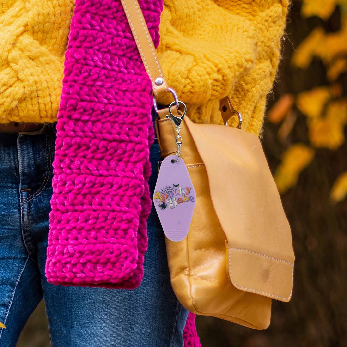 Lilac motel-style keychain with "Spooky Babe" in orange and purple, featuring a spider and web, clipped to a tan leather purse, beside a bright pink scarf and yellow sweater.