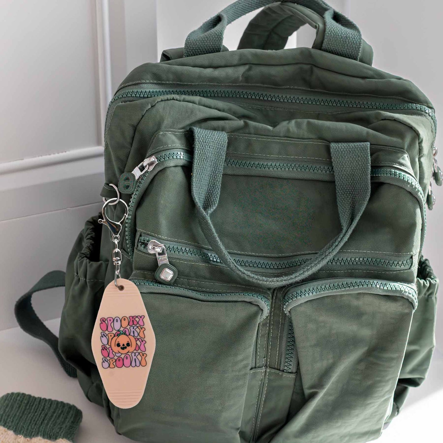 Peach colored motel-style keychain with 'SPOOKY' text and an orange pumpkin with a cute face, attached to a green backpack.