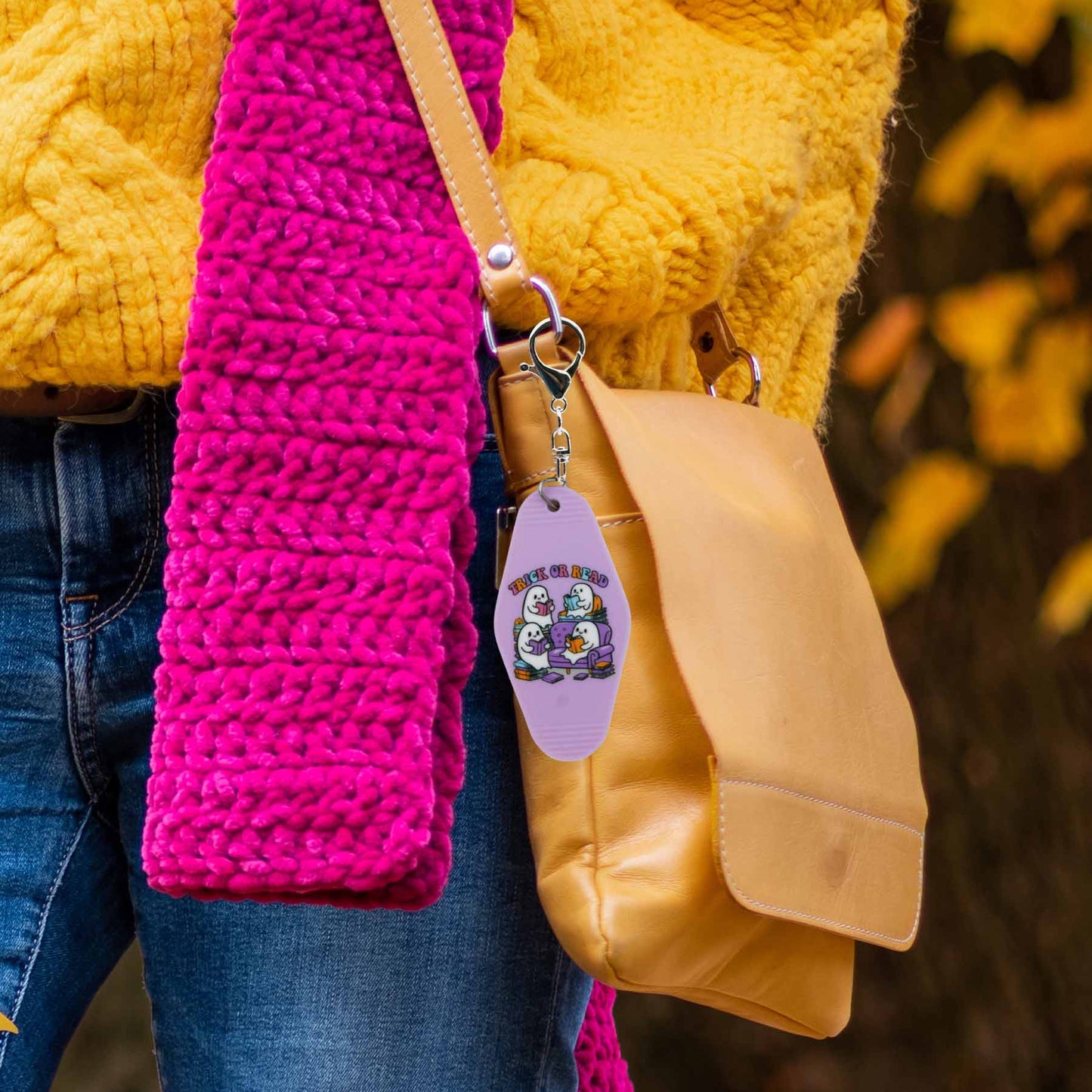 Lavender "Trick or Read" keychain with cute ghost characters reading on a couch, attached to a tan leather bag strap, next to a pink crocheted scarf, yellow sweater, and blue jeans.