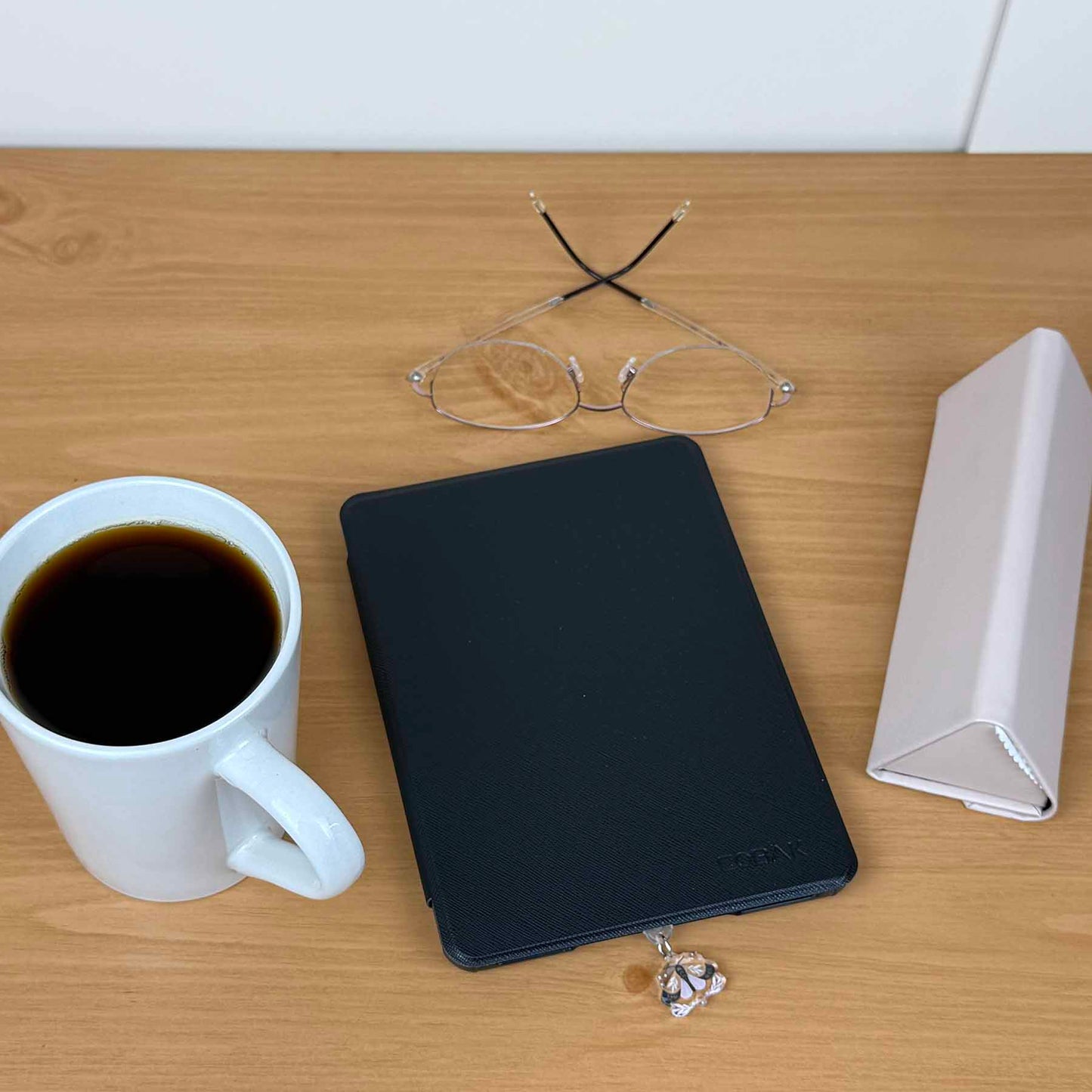 Moth Kindle Charm USB-C dust plug attached to a black Kindle on a wooden table with coffee and glasses.