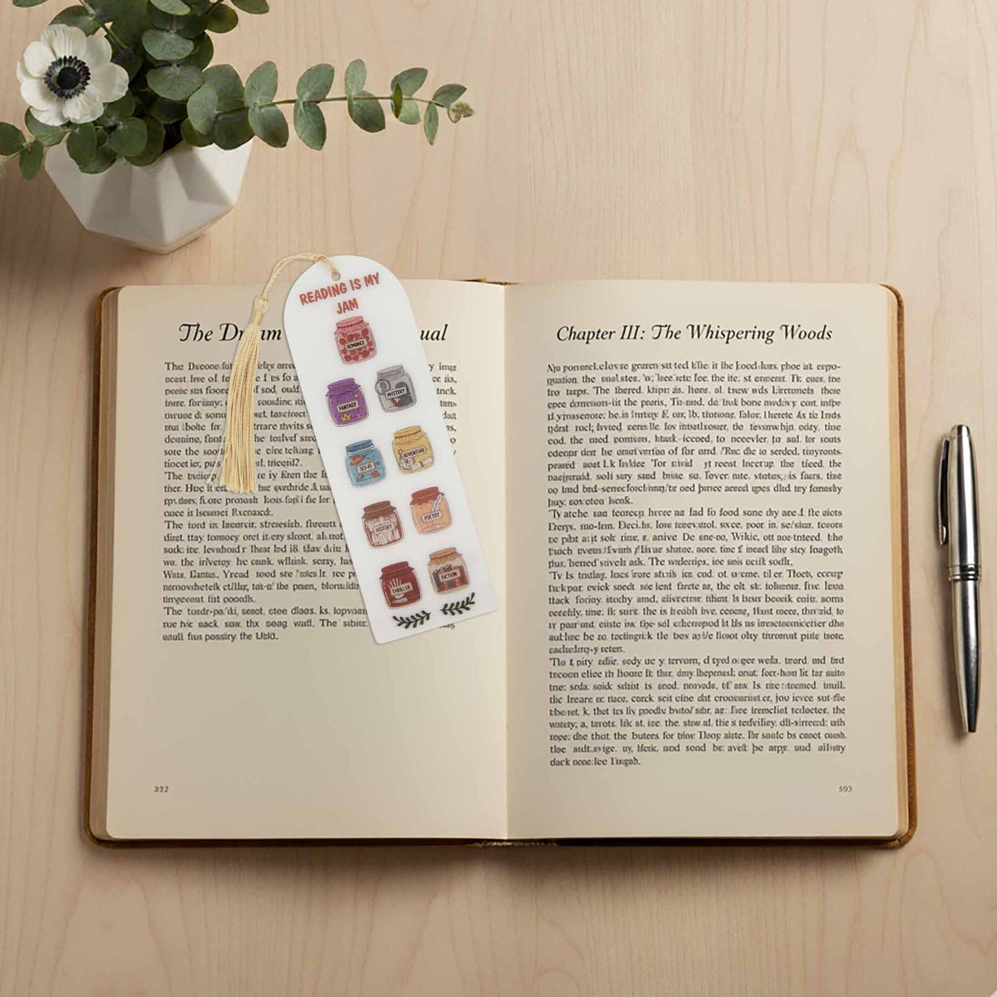 Bookmark with 'Reading Is My Jam' text and various genre-themed jam jar illustrations, placed in an open book with text on a light wood surface.