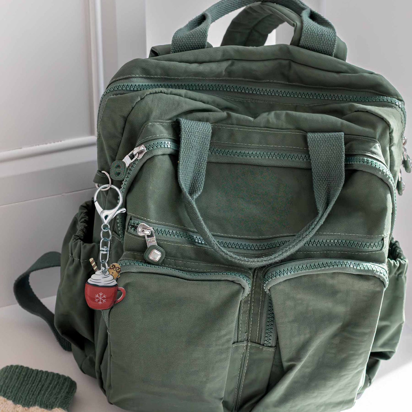 Winter hot cocoa bag charm keychain with snowflake design attached to a green backpack, a green backpack