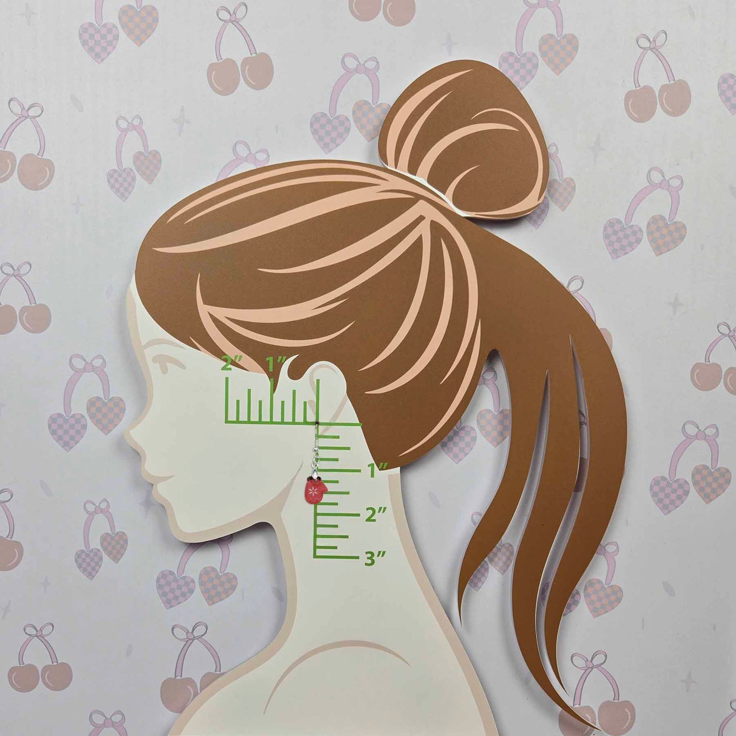 Single red mitten earring with white snowflake detail, dangling from a model's ear. Green ruler markings show the earring is approximately 1.5 inches long.
