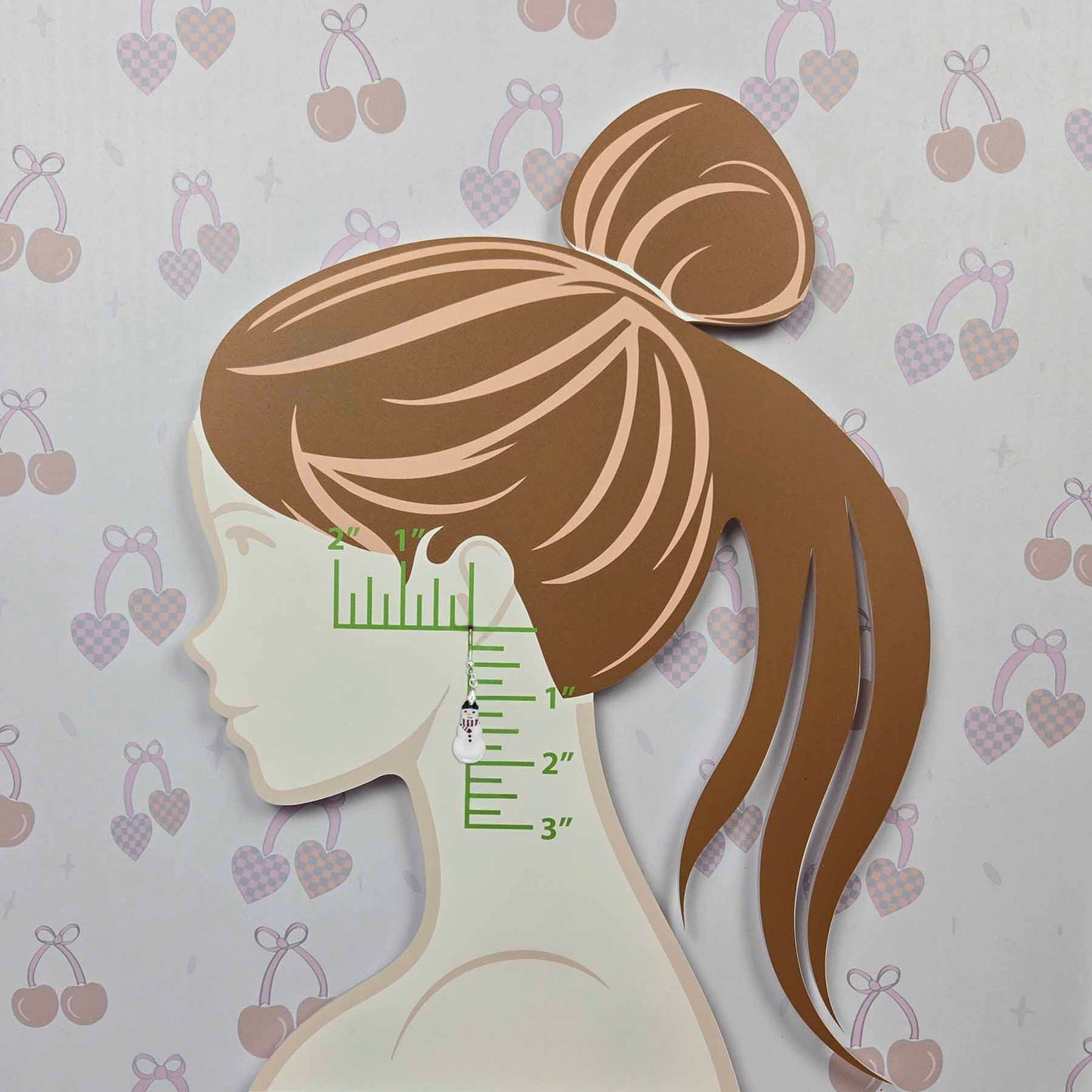 Snowman dangle earring on a display board, showing its size against an inch ruler. The white snowman has a black top hat and red scarf. The board is shaped like a woman's profile with brown hair, on a light gray background with cherry and heart patterns.