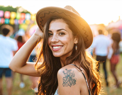 Smiling woman in cowboy hat and unique drink cup earrings at outdoor festival, sun flare, shoulder tattoo.