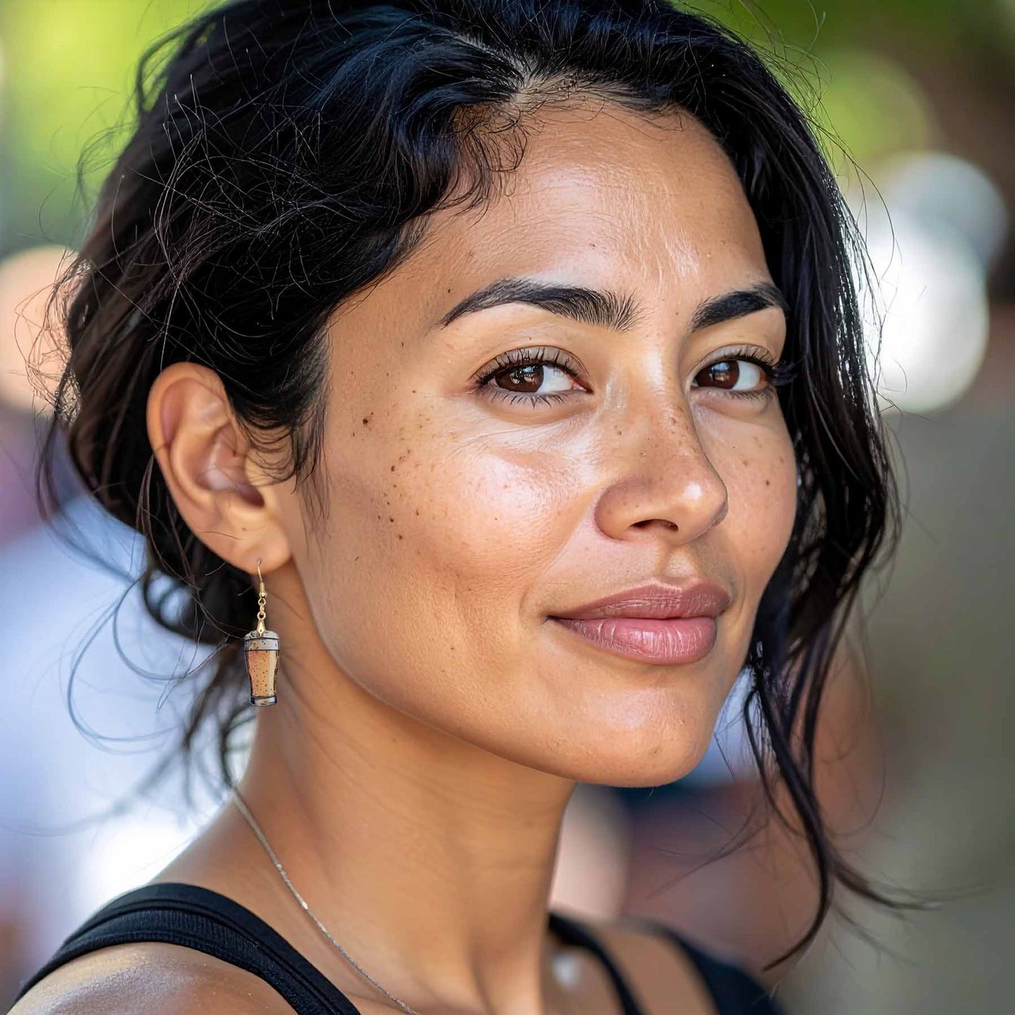 Close-up of a woman with dark hair and brown eyes, wearing a black tank top and unique beer glass dangle earrings. Her face has freckles, and she is subtly smiling.