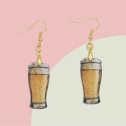 A pair of dangle earrings featuring miniature beer glasses filled with golden ale and foamy heads, suspended from gold-colored hooks.