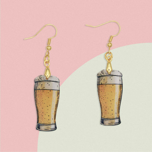 A pair of dangle earrings featuring miniature beer glasses filled with golden ale and foamy heads, suspended from gold-colored hooks.
