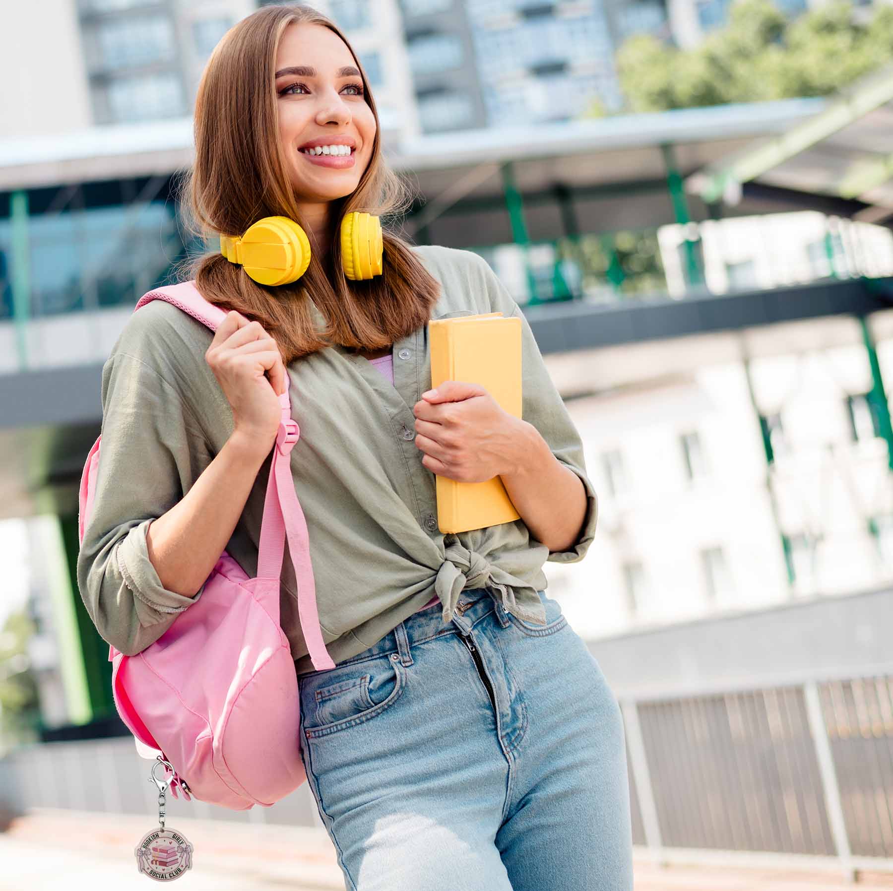 Smiling girl with yellow headphones, a pink backpack with a 'Bookish Girls Social Club' keychain, and a yellow book. She wears a sage green tied shirt and blue jeans, looking right.