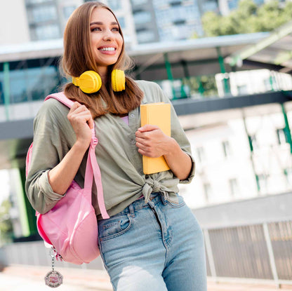 Smiling girl with yellow headphones, a pink backpack with a 'Bookish Girls Social Club' keychain, and a yellow book. She wears a sage green tied shirt and blue jeans, looking right.