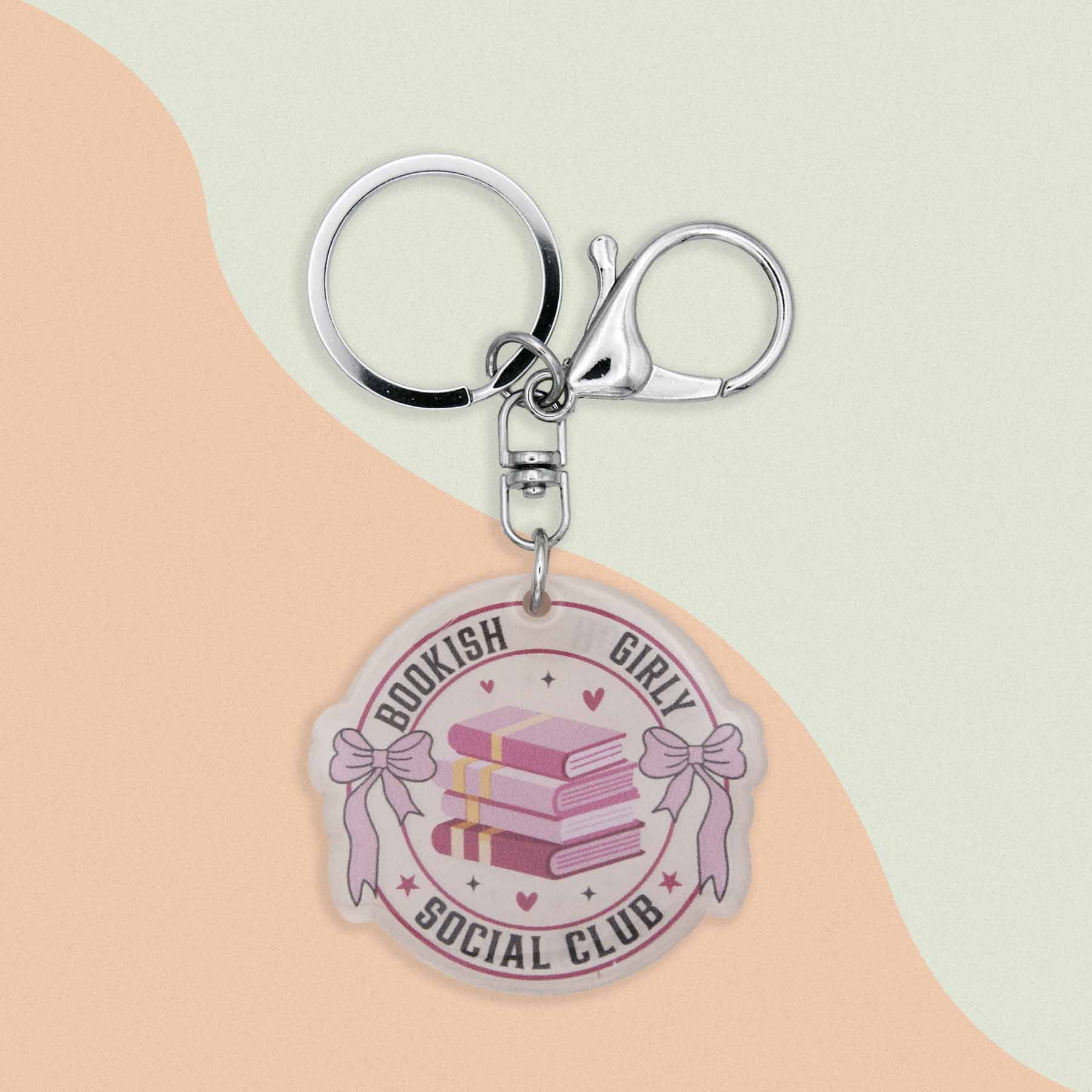 Acrylic keychain with a round charm featuring three pink books, pink bows, and text that says 'Bookish Girly Social Club' with a silver keyring and clasp.