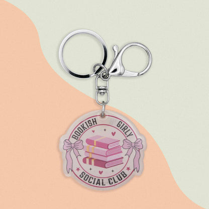 Acrylic keychain with a round charm featuring three pink books, pink bows, and text that says 'Bookish Girly Social Club' with a silver keyring and clasp.