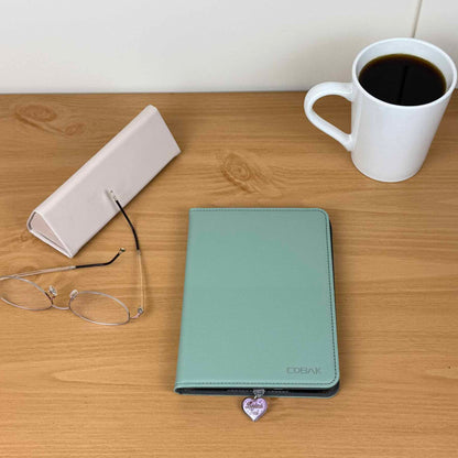 Heart-shaped "Bookish Soul" Kindle charm attached to a light green Kindle cover on a wooden desk with glasses and coffee.