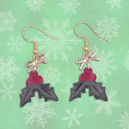 A pair of Christmas holly leaf earrings with red berries, gold bows, and hooks, set against a green snowflake background.