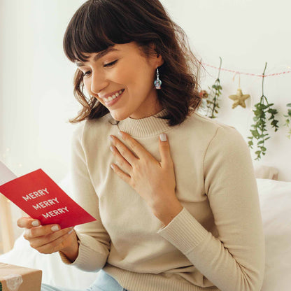 Smiling woman wearing a holiday snow globe earring, holding a red 'Merry' Christmas card, in a festive room.
