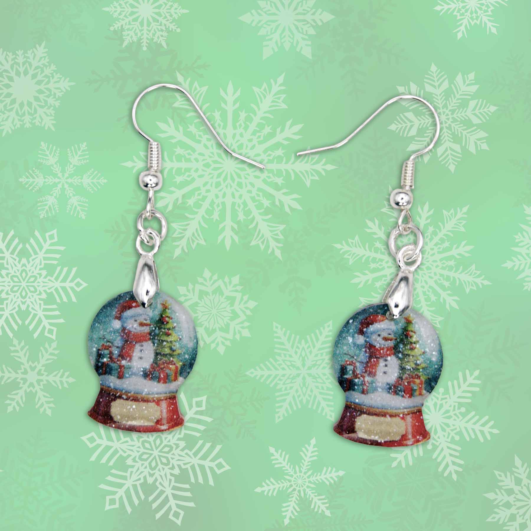 A pair of dangle earrings featuring miniature snow globe charms. Each charm depicts a snowman in a red hat and scarf, standing next to a decorated Christmas tree and gifts, with falling snow inside the globe. The base of the snow globe is red. The earrings have silver-toned hooks and hang against a light green background with white snowflake patterns.