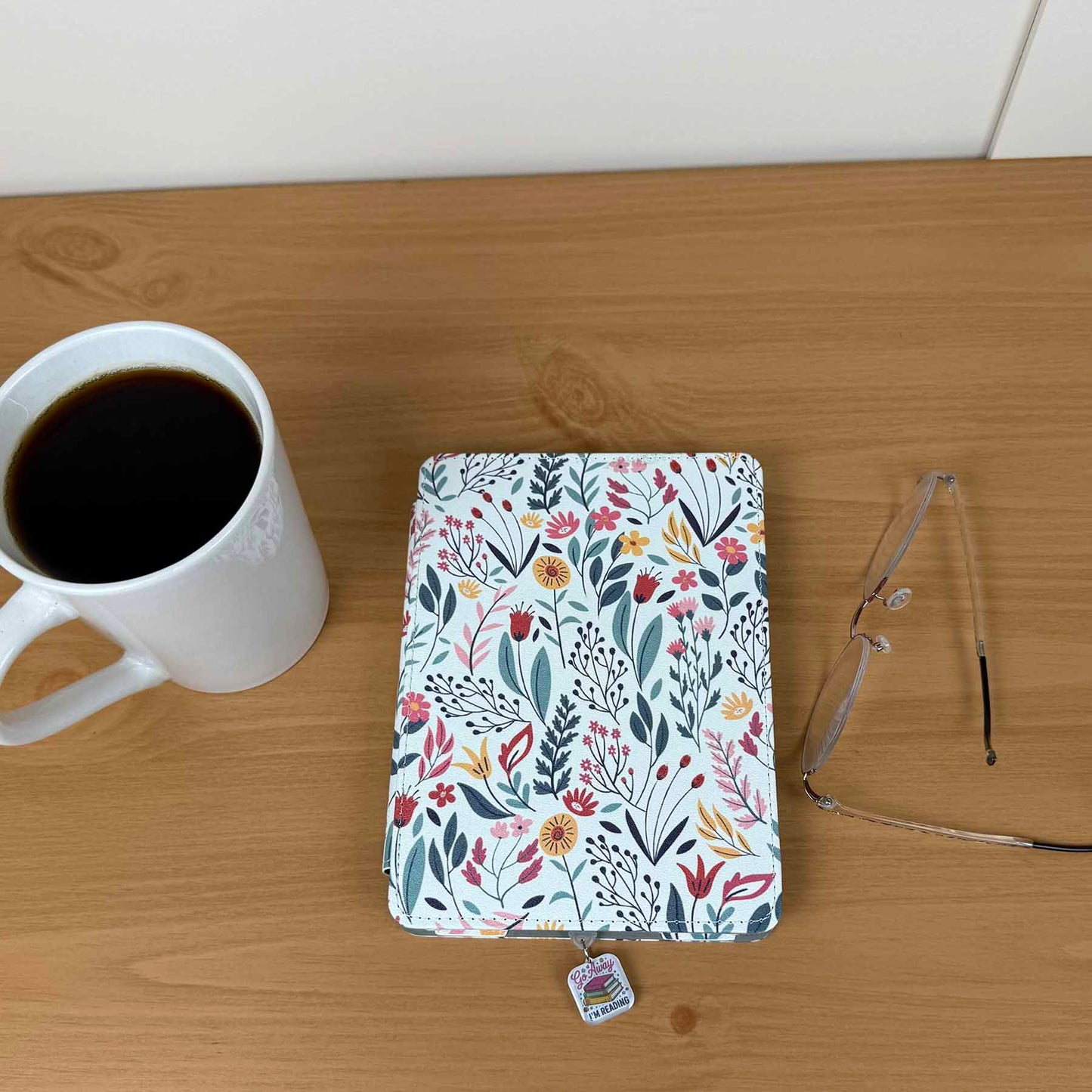 Go Away I'm Reading Kindle Charm attached to a floral Kindle cover next to a coffee cup and glasses