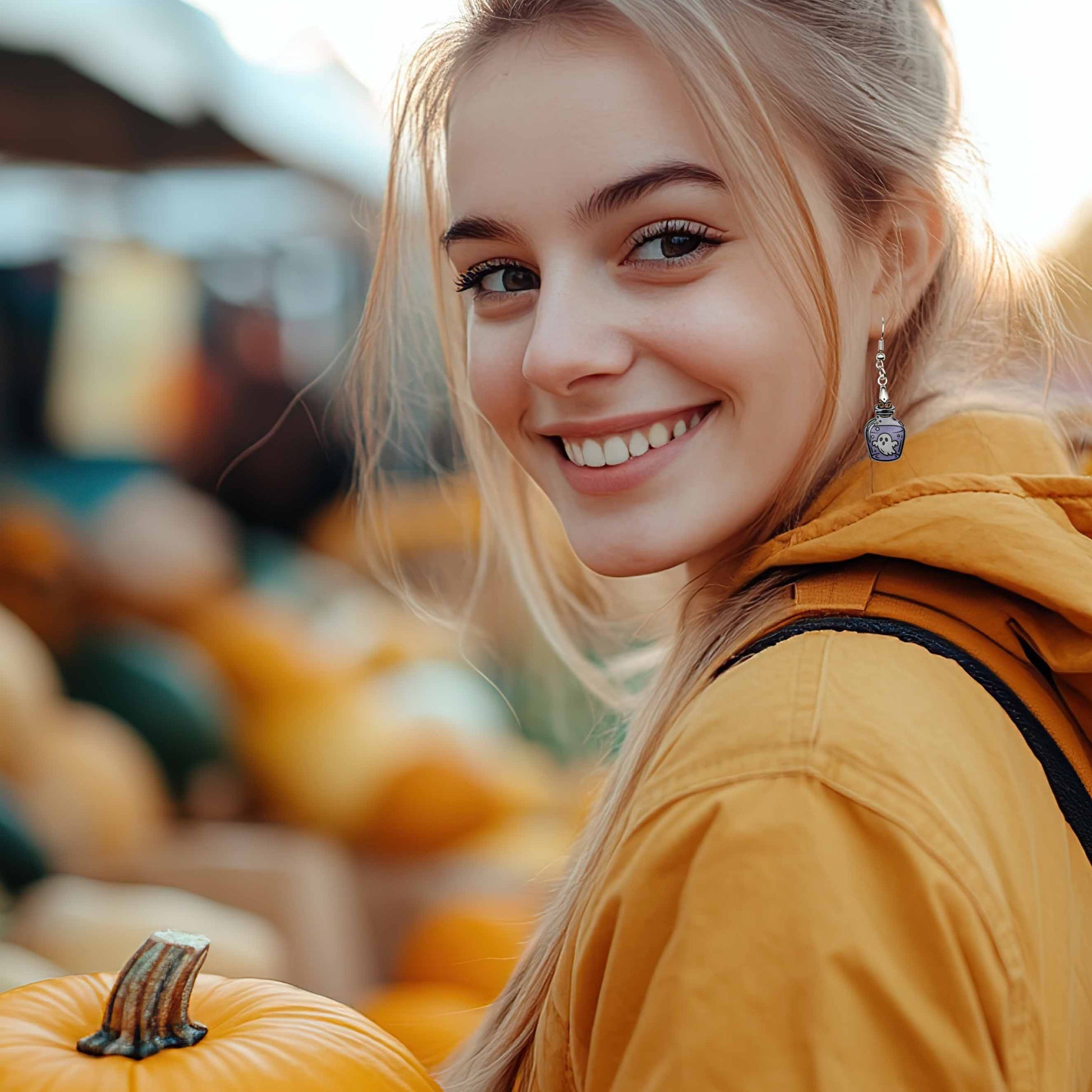 Blonde woman in yellow jacket smiling, wearing a Halloween ghost in bottle potion dangle earring. Holding a pumpkin at an outdoor market.