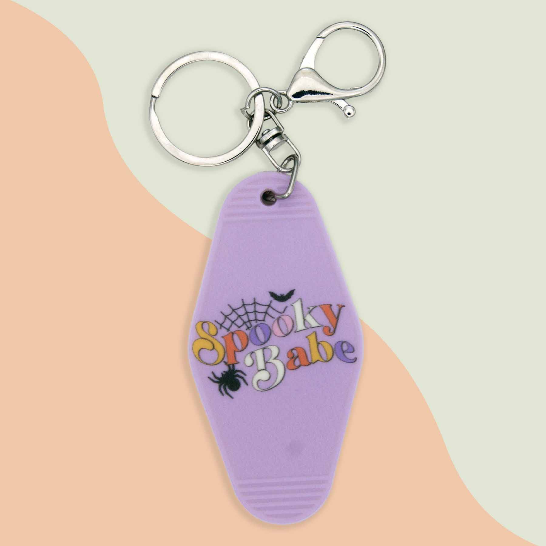 Spooky Babe keychain with a purple hotel-style key tag, featuring colorful text, a spiderweb, a spider, and a bat design, attached to a silver keyring and clasp.