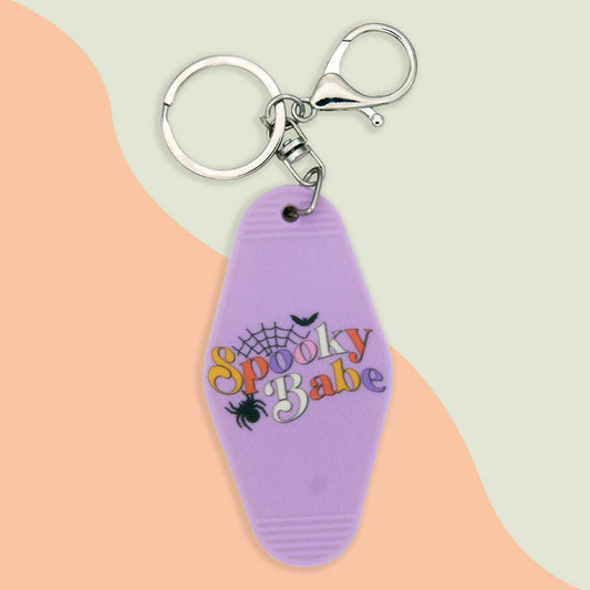 Spooky Babe keychain with a purple hotel-style key tag, featuring colorful text, a spiderweb, a spider, and a bat design, attached to a silver keyring and clasp.