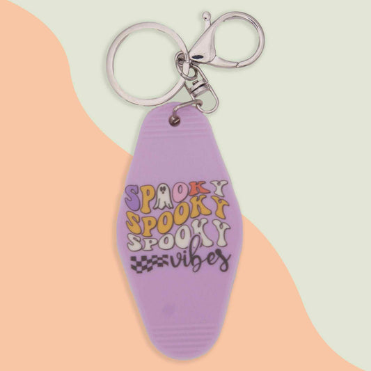 Lavender motel-style keychain with 'Spooky Spooky Spooky Vibes' text in purple, orange, and black, featuring ghost and checkered flag details.