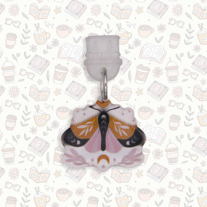 Moth Kindle Charm, a USB-C dust plug, with intricate moth design in pink, black, and gold colors