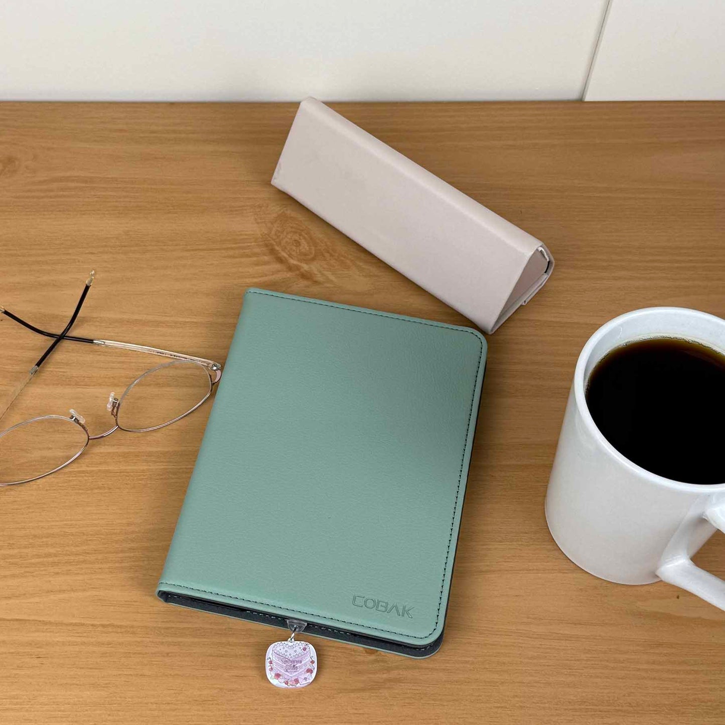 Romance Girlie Cake Kindle charm on a green Kindle cover, with glasses, coffee mug, and white case on a wooden desk