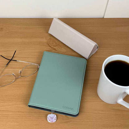 Romance Girlie Cake Kindle charm on a green Kindle cover, with glasses, coffee mug, and white case on a wooden desk