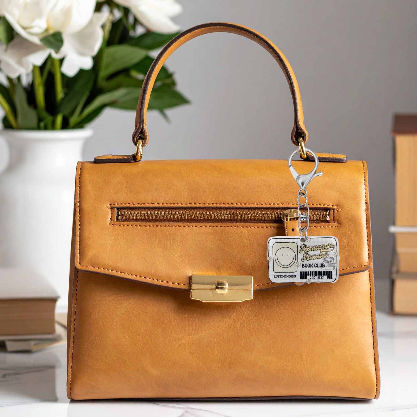 Romance Reader Book Club library card keychain hanging from a brown leather handbag. The keychain is white with a smiling face, text "Romance Reader BOOK CLUB," "LIFETIME MEMBER," and a barcode.