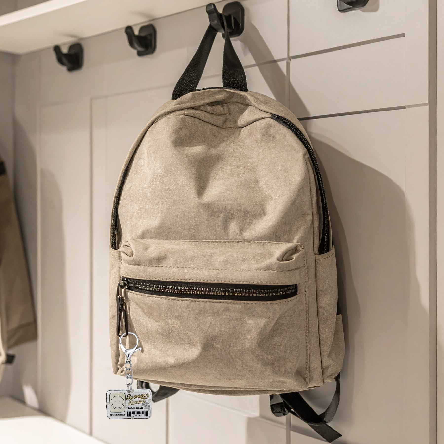 A tan or light beige textured backpack with black zippers and straps, hanging on a black hook on a white wall. A 'Romance Reader Book Club Lifetime Member' keychain with a smiley face is clipped to a zipper pull.