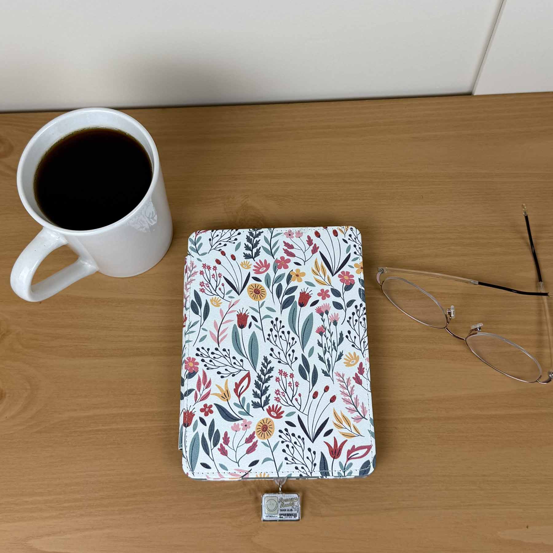Romance Reader Book Club Kindle Charm attached to a floral e-reader next to a coffee mug and glasses on wood.