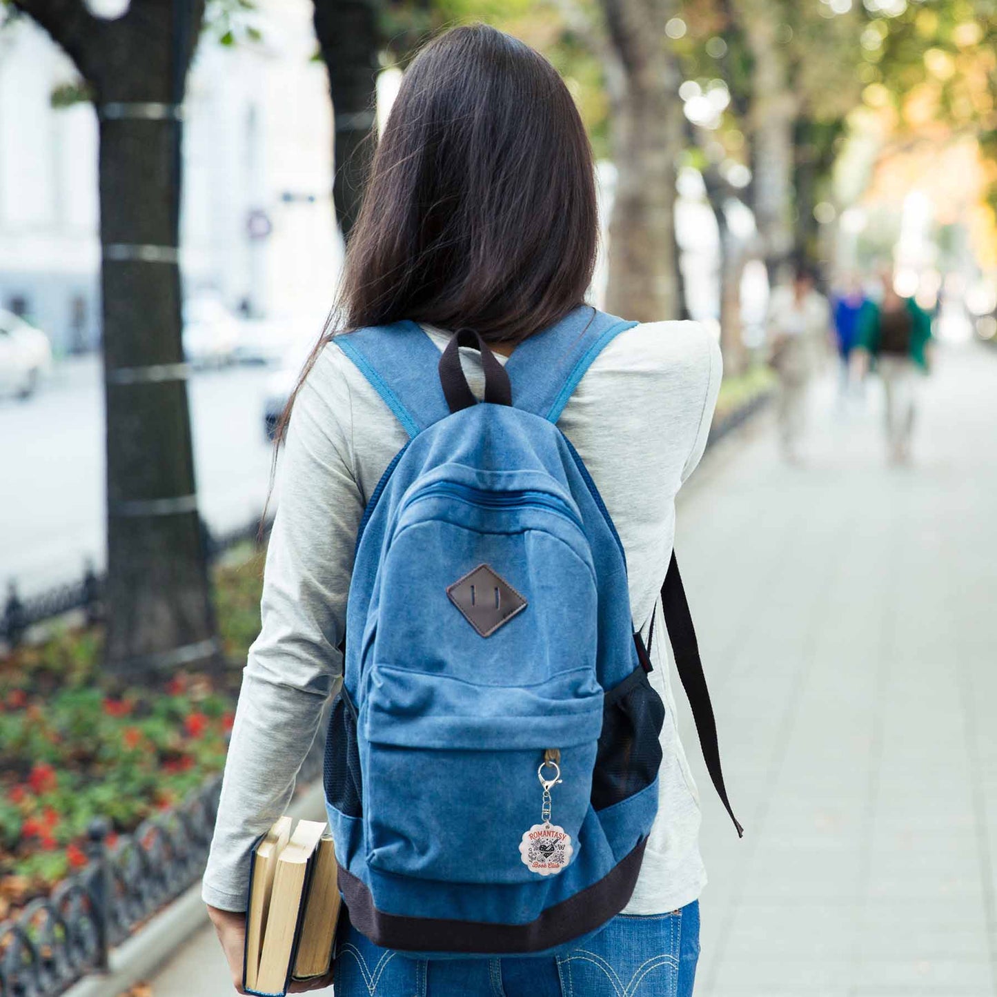 Young woman with long dark hair, wearing a grey shirt and jeans, walking away, carrying a blue canvas backpack with a "ROMANTASY Book Club" keychain, and holding books in her left hand, on a sidewalk next to trees.