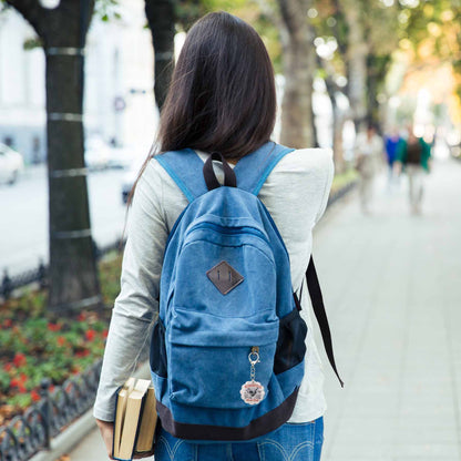 Young woman with long dark hair, wearing a grey shirt and jeans, walking away, carrying a blue canvas backpack with a "ROMANTASY Book Club" keychain, and holding books in her left hand, on a sidewalk next to trees.