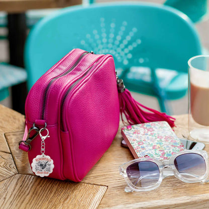 Pink textured leather crossbody bag with dual zippers, a matching tassel, and a round 'Romantasy Book Club' charm, resting on a wooden table next to clear-framed sunglasses, a floral notebook, and a glass.