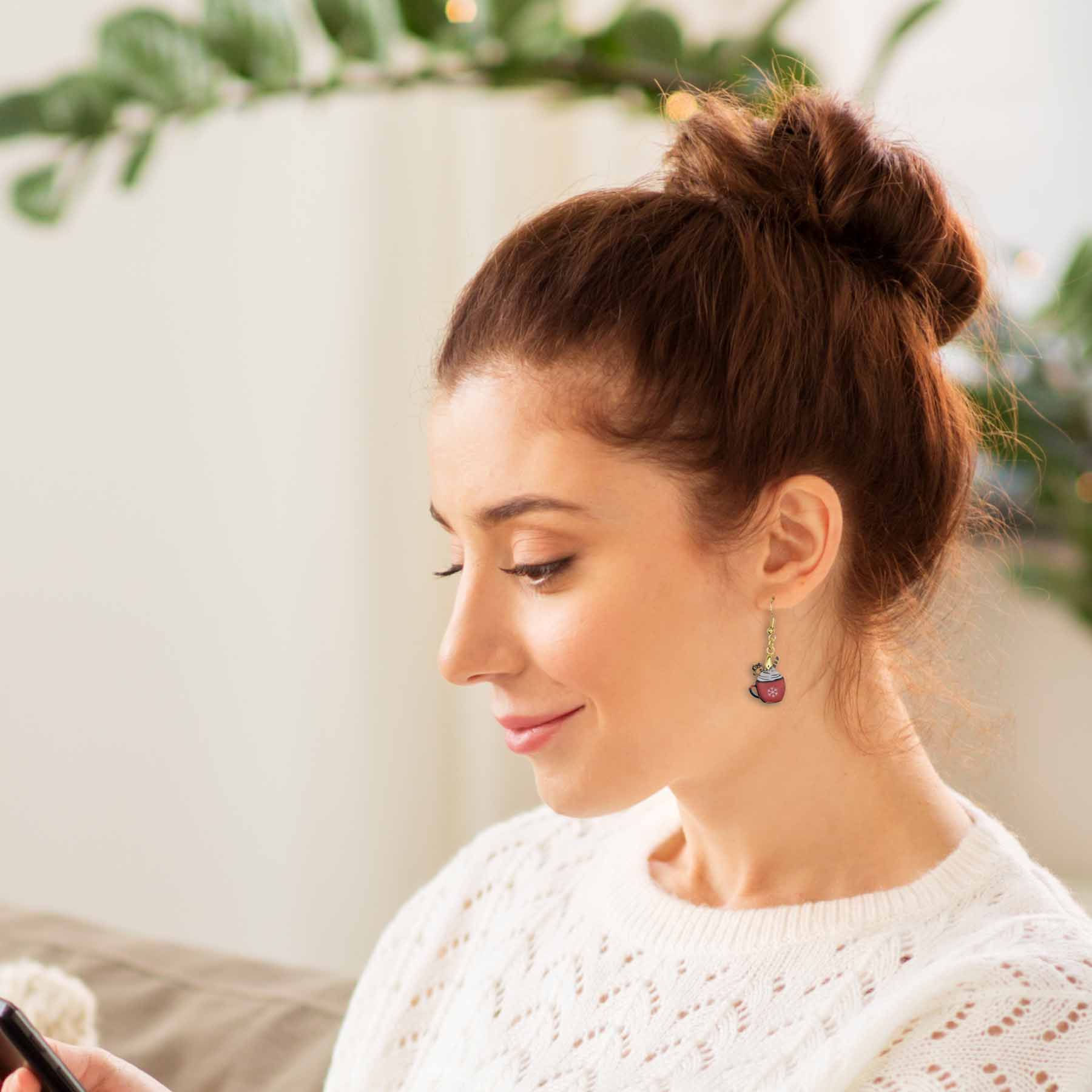 Woman wearing gold dangle earrings with red hot cocoa cup charms, featuring white whipped cream and snowflakes, paired with a white knit sweater.