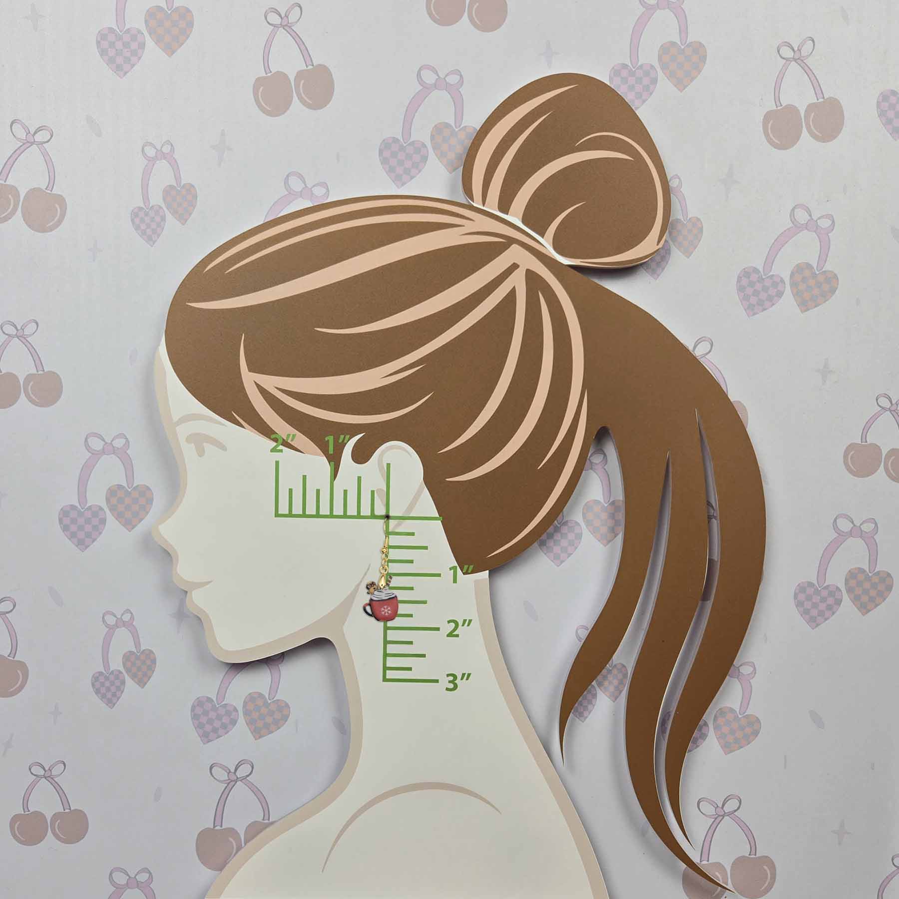 Single red hot cocoa mug dangle earring with white whipped cream, snowflake, and gold gingerbread man charm, shown on an ear model with a ruler indicating 1.5-inch length.