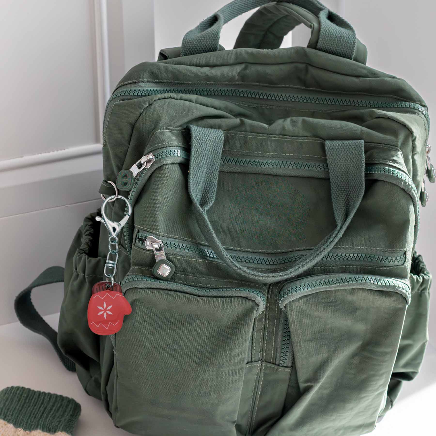 Red acrylic winter mitten bag charm keychain with a snowflake design, attached to a green backpack.