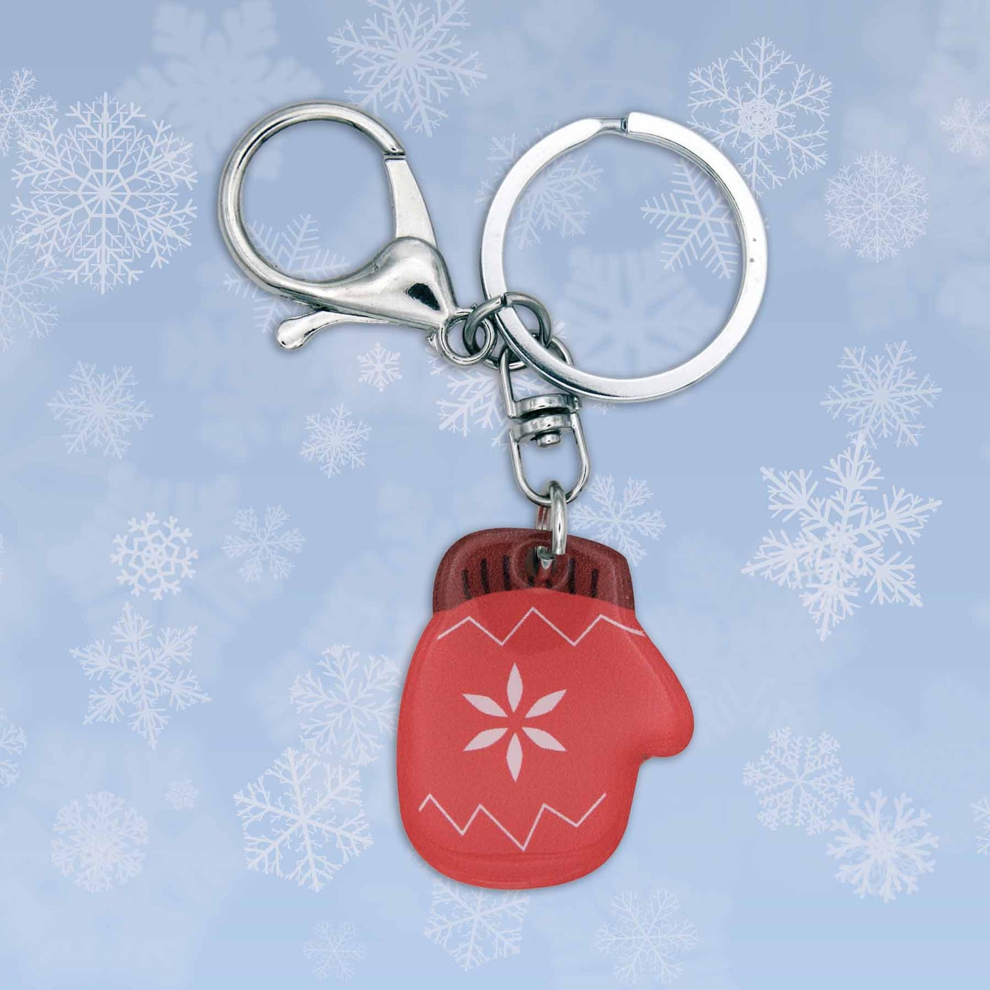 Red acrylic winter mitten bag charm keychain with a snowflake design, silver clasp, and keyring on a snowy background.