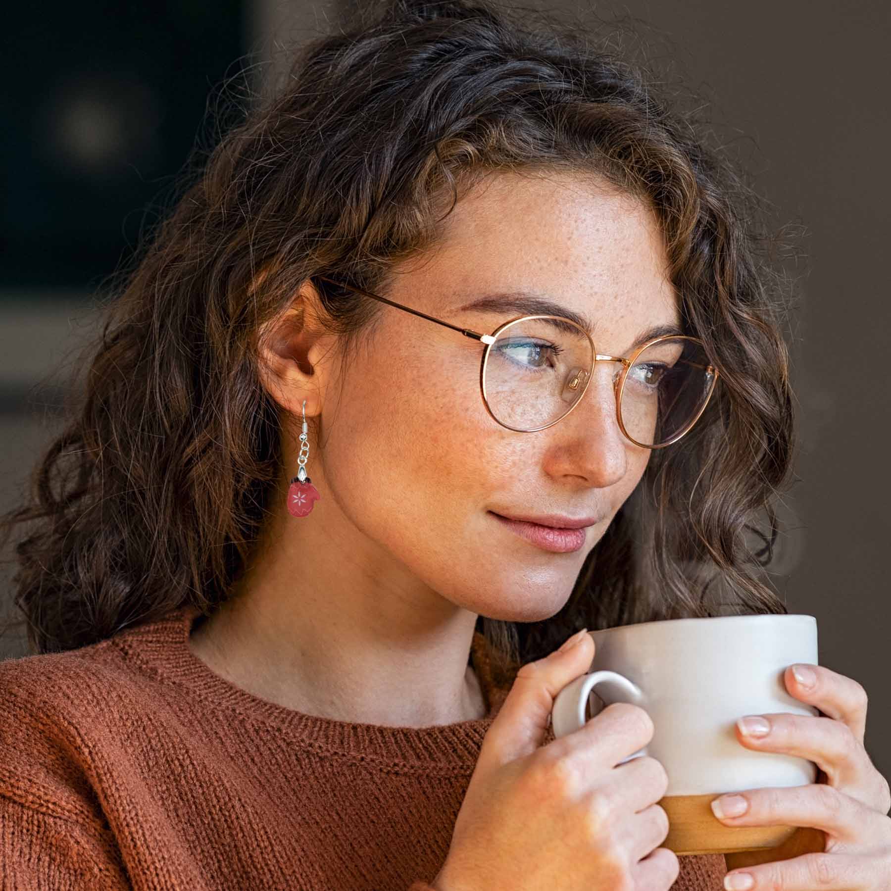 Woman with curly brown hair wearing glasses, a rust-colored sweater, and dangling red mitten earrings with white snowflake detail, holding a white and wood-toned mug.