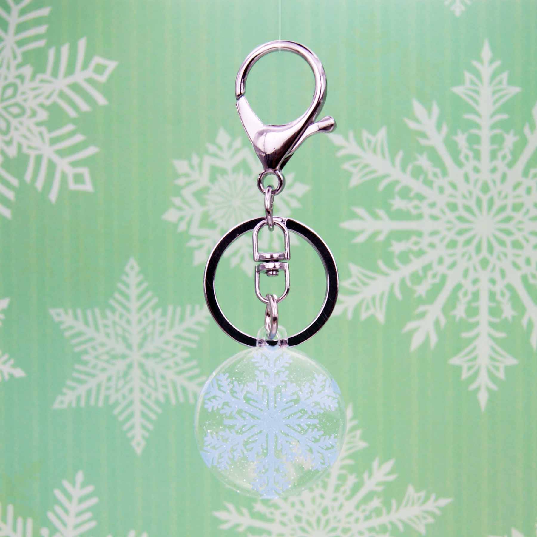 Sparkly clear acrylic snowflake bag charm keychain with silver clasp and keyring against a green snowy background.
