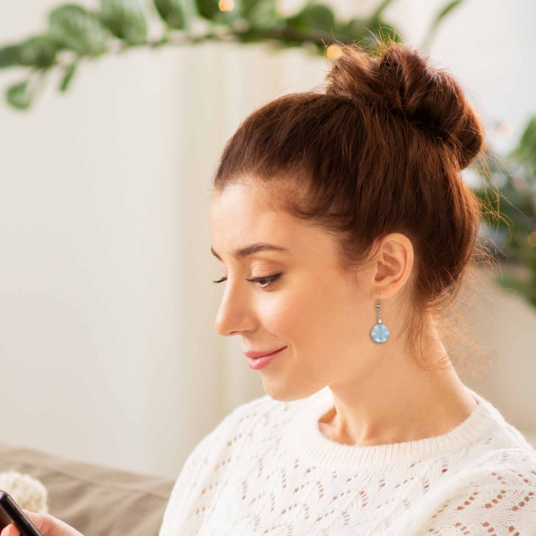 Close-up of a woman wearing light blue circular snowflake drop earrings, paired with a white knit sweater. Her auburn hair is in a bun.