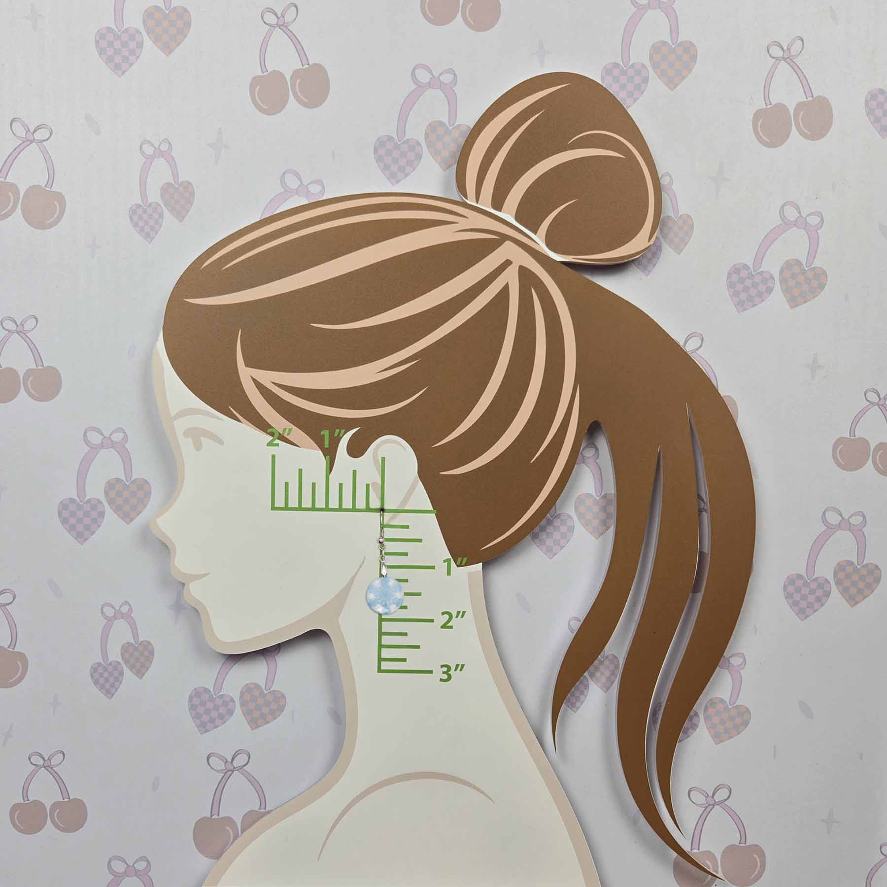 Single blue and white cloudy sky patterned dangle earring with silver hook, shown on a mannequin head with a ruler marking 1-3 inches for size reference. The background has a light pattern of cherries and hearts.